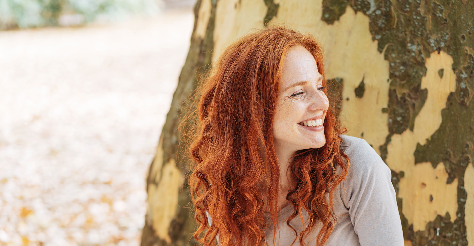 Smiling young woman leaning against the trunk of a tree with peeling bark looking to the side with a beaming smile of pleasure