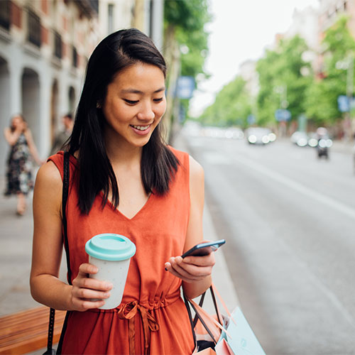 woman stands on sidewalk carrying coffee in one hand and looking at her phone
