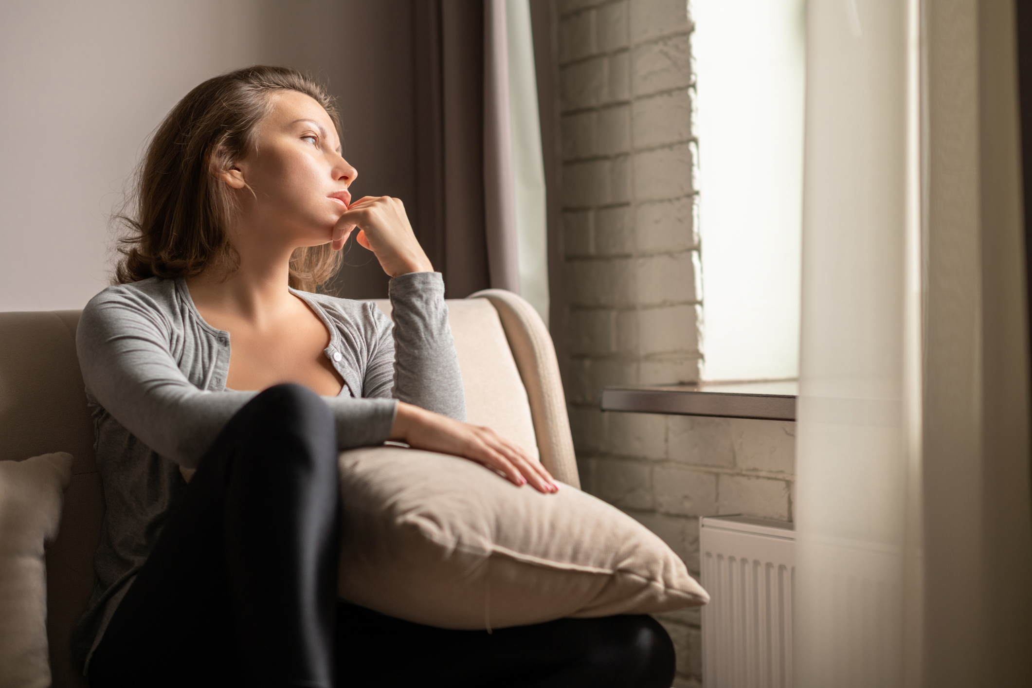 Portrait of lonely sad young woman sitting near window of apartments.